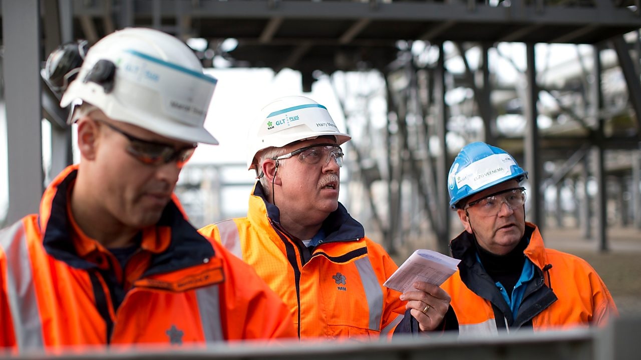 Medium foto van drie medewerkers in oranje jassen, met witte helmen en veiligheidsbrillen
