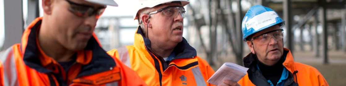 Medium foto van drie medewerkers in oranje jassen, met witte helmen en veiligheidsbrillen