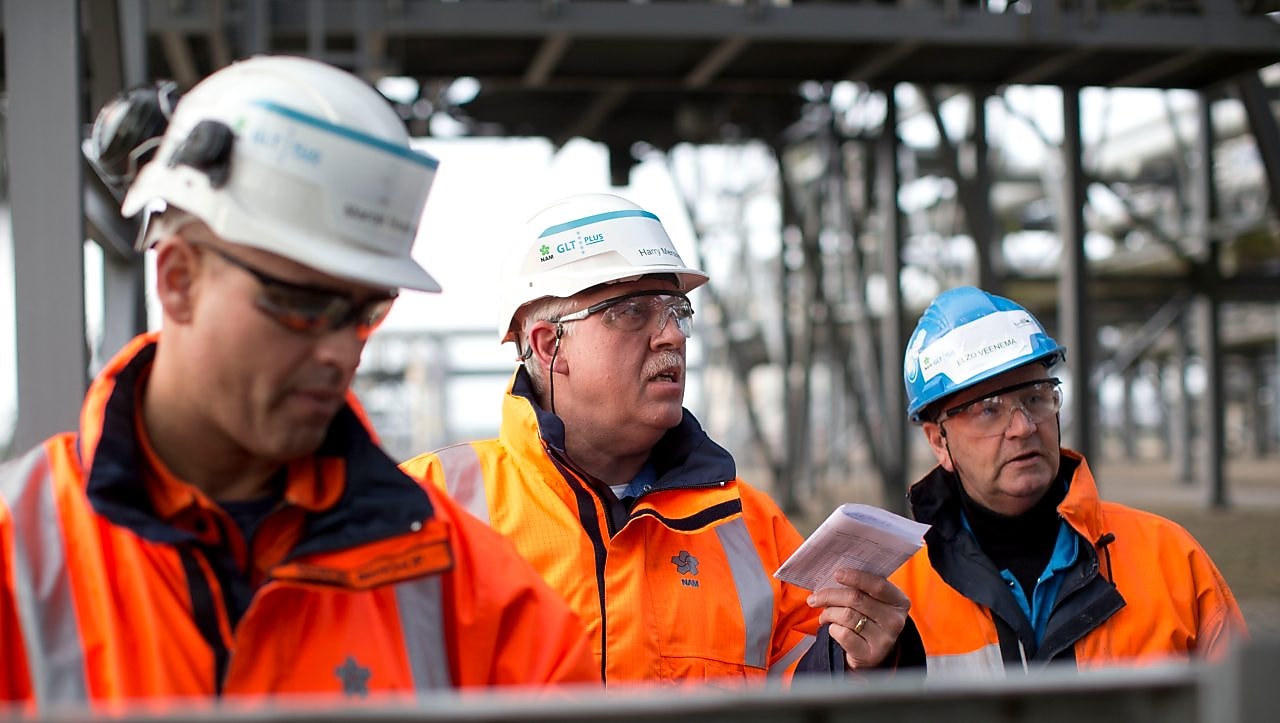 Medium foto van drie medewerkers in oranje jassen, met witte helmen en veiligheidsbrillen