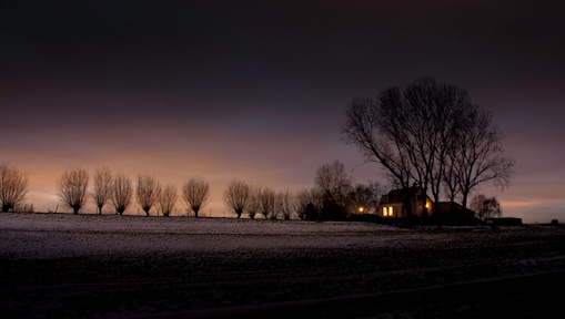 Winterlandschap in de schemer met kale bomen en een huis waarin lichtbrand
