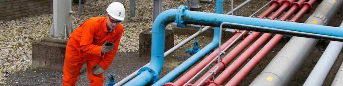 Medewerker in oranje overall met portofoon naast grijze, blauwe en rode buizen