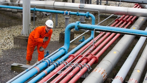 Medewerker in oranje overall met portofoon naast grijze, blauwe en rode buizen