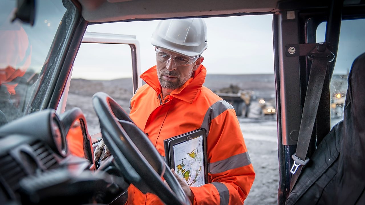 Medewerker in oranje overall met tablet onder arm gezien vanuit een auto